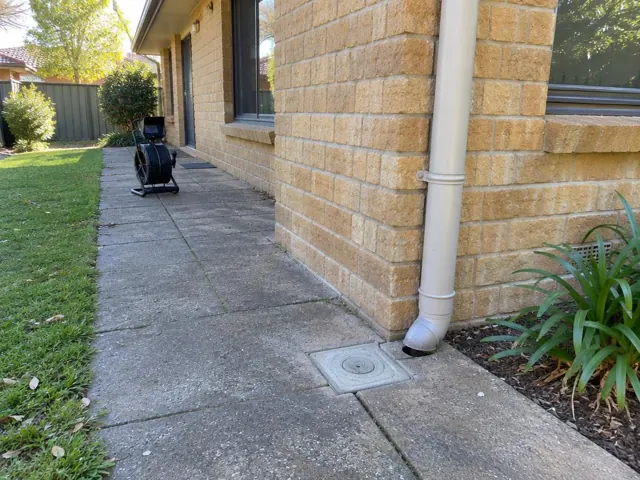Capped drain inspection cover beside a house wall and downpipe on a paved side path in Kings Park