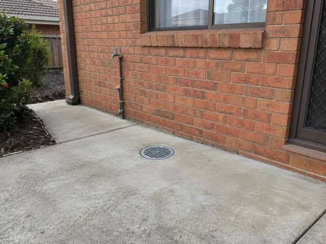 Concrete patio beside a brick house with a round drain grate and wall pipework in Blacktown