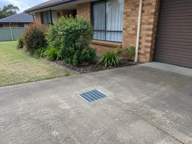 Concrete driveway with a square stormwater grate beside a brick house and garden in Marayong
